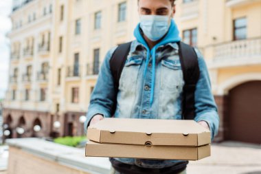 Tıbbi maskeli teslimatçının seçici odak noktası elinde pizza kutuları ve arka planda şehir caddesi. 