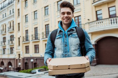 Pizza kutuları taşıyan yakışıklı kurye ve şehir sokağındaki kameraya gülümsüyor. 
