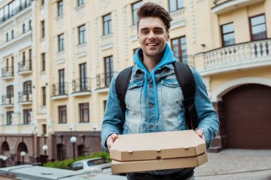 Handsome courier holding pizza boxes and smiling at camera on urban street  clipart