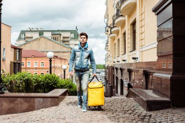 Handsome delivery man holding thermo bag while walking near buildings on paving stone walkway clipart