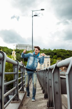 Selective focus of positive man in earphones dancing on bridge with urban street and cloudy sky at background  clipart