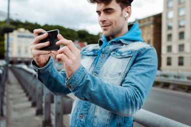 Kulaklıklı yakışıklı bir adamın akıllı telefonuyla şehir sokaklarında fotoğraf çekmesi seçici bir odak noktası. 