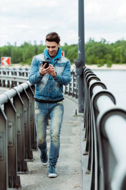 Köprüde yürürken akıllı telefon kullanan gülümseyen adamın seçici odağı 