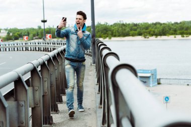 Köprüde akıllı telefonla selfie çekerken barış işareti gösteren neşeli adamın seçici odağı 