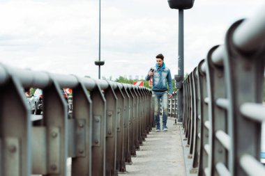 Selective focus of handsome man using smartphone while walking on bridge walkway clipart