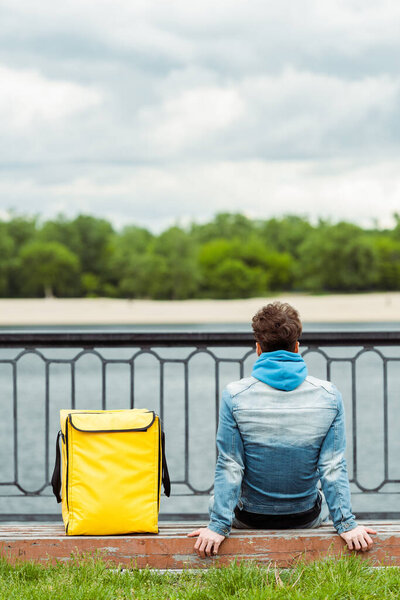 Back view of courier sitting on bench near thermo bag on urban street 