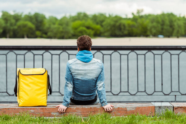Back view of delivery man sitting near thermal bag on bench on promenade