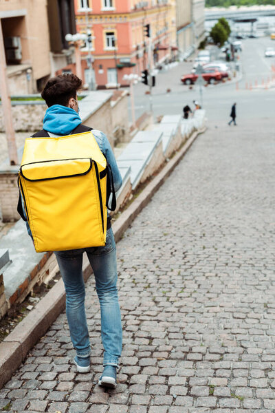 Back view of courier in medical mask with thermo backpack walking near on urban street