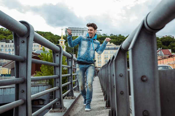 Selective focus of cheerful man in earphones showing yeah gesture while walking on bridge 