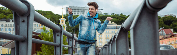 Panoramic crop of positive man in earphones showing yeah gesture while walking on bridge 