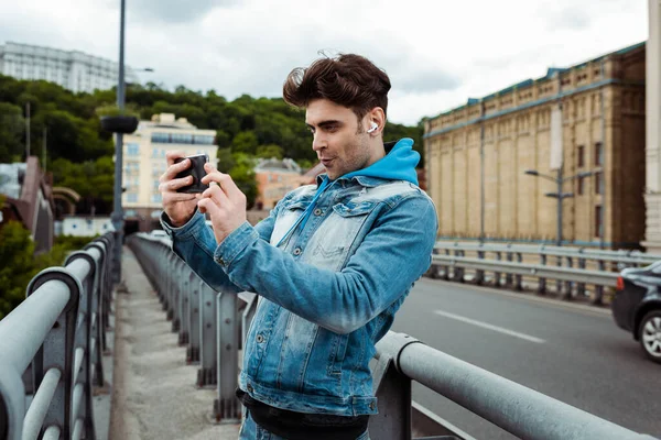 Handsome man in earphones taking picture with smartphone on urban street