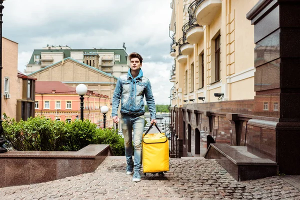 Handsome delivery man holding thermo bag while walking near buildings on paving stone walkway — Stock Image Handsome Delivery Man Holding Thermo Bag While Walking Buildings Paving Stock Image