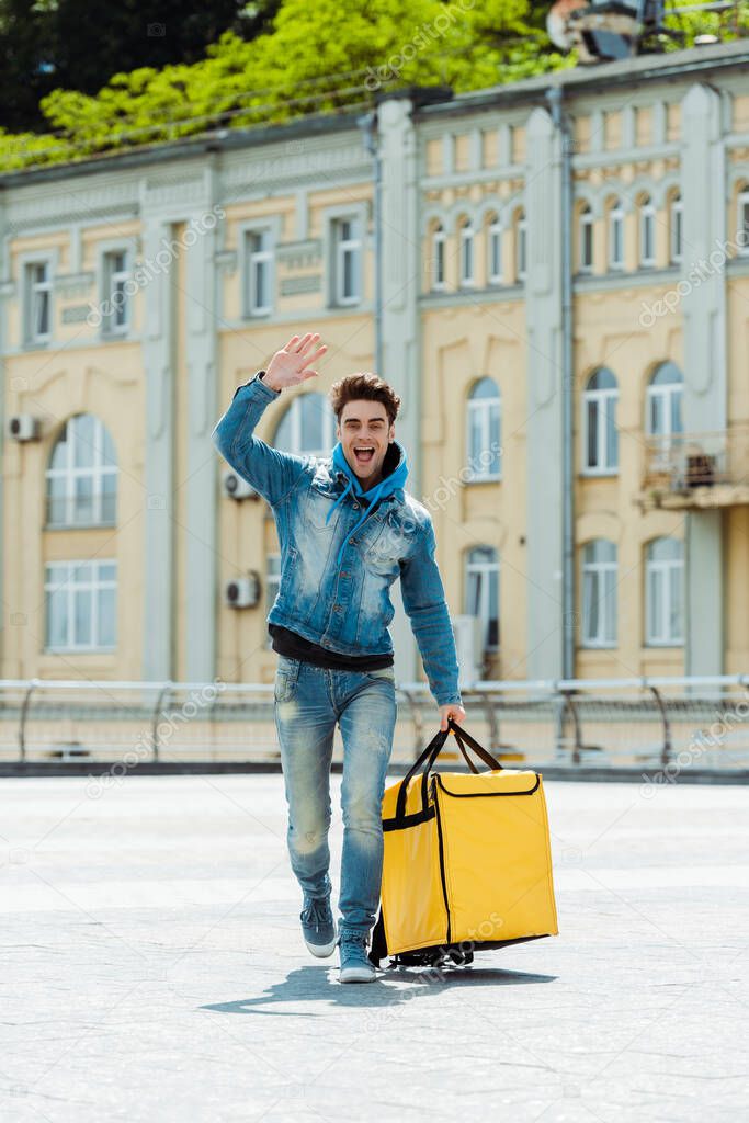 Positive courier with thermo bag waving hand on urban street 