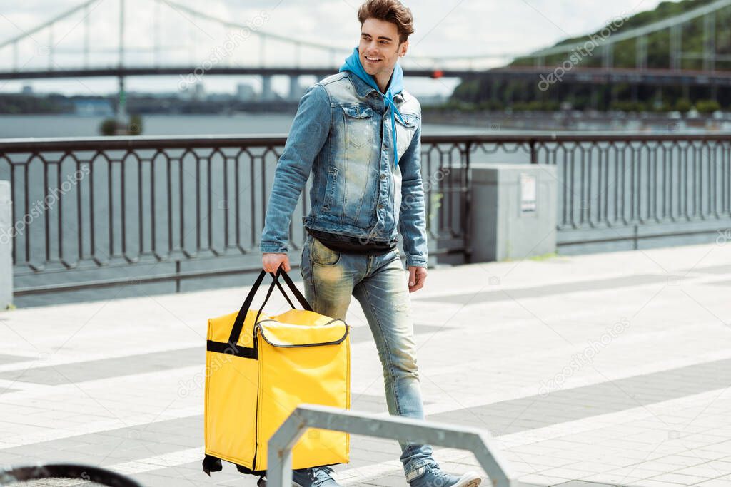 Selective focus of smiling courier with thermo bag walking on urban street 