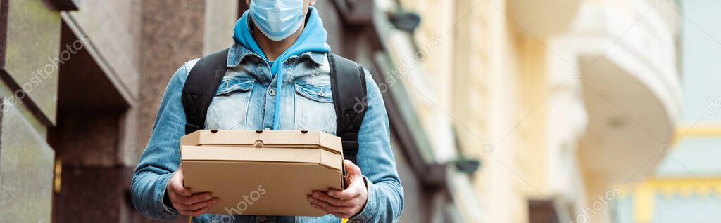 Cropped view of courier in medical mask holding pizza boxes on urban street, panoramic shot 