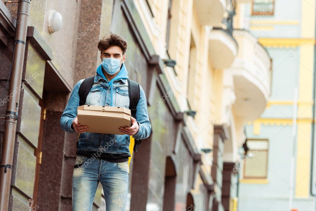 Delivery man in medical mask holding pizza boxes near facade of building 