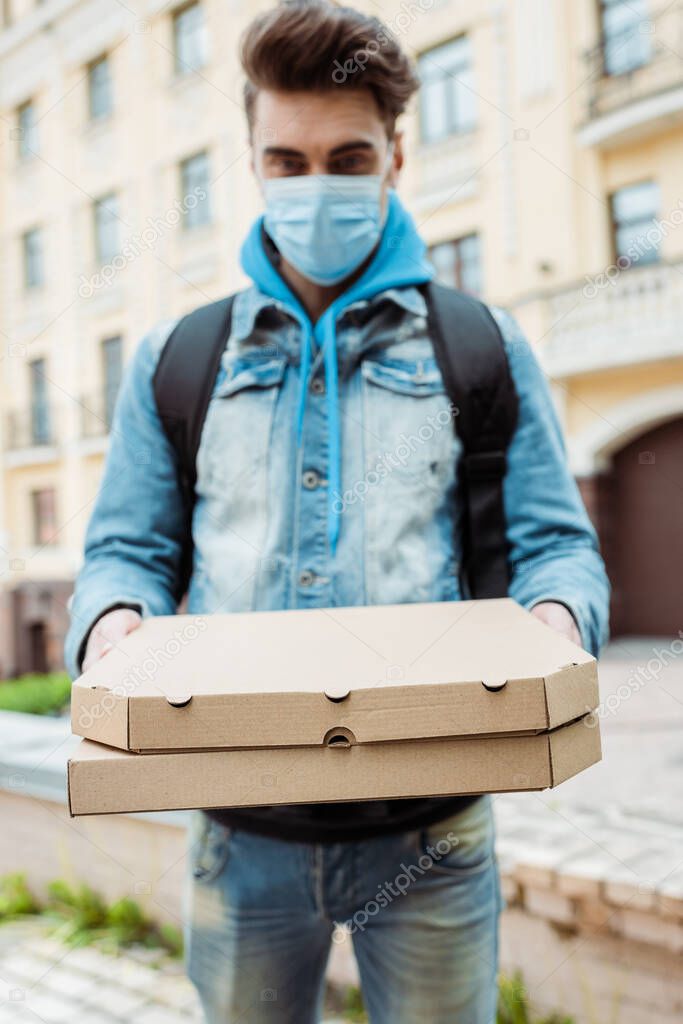 Selective focus of courier in medical mask holding cardboard pizza boxes on urban street 