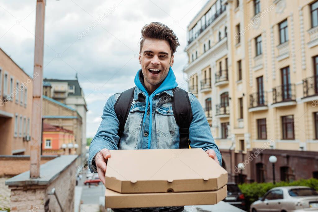 Selective focus of cheerful courier holding pizza boxes with urban street at background 