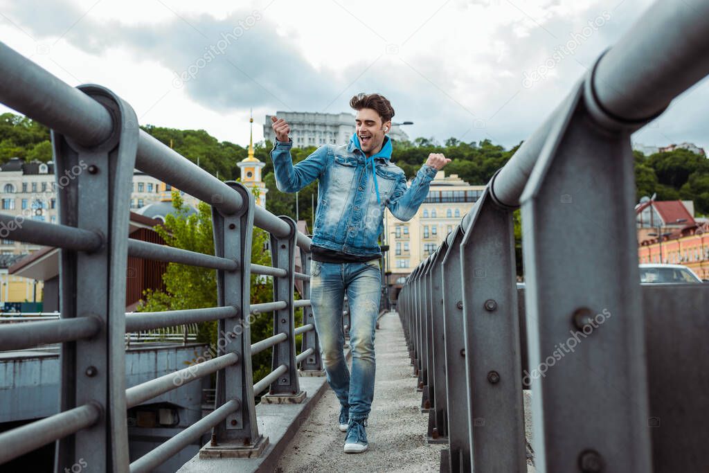 Selective focus of cheerful man in earphones showing yeah gesture while walking on bridge 