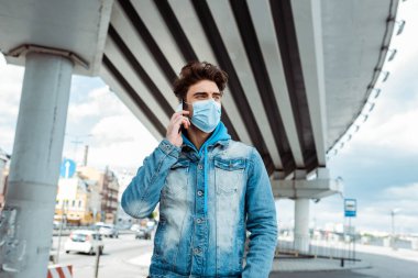 Tıbbi maskeli bir adam şehir caddesinde akıllı telefondan konuşuyor. 