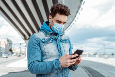 Tıp maskeli genç adam şehir caddesinde akıllı telefon kullanıyor.