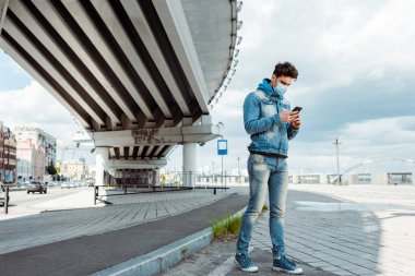 Tıbbi maskeli ve kot ceketli bir adam köprünün yanındaki şehir sokağında akıllı telefon kullanıyor. 