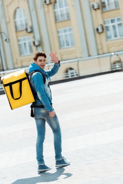 Şehir caddesinde el sallayan termo sırt çantalı mutlu kuryenin arka görüntüsü