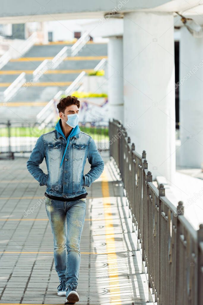 Young man in medical mask with hands in pockets of jacket walking on urban street 