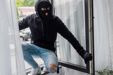 Selective focus of robber in balaclava standing on open window  clipart