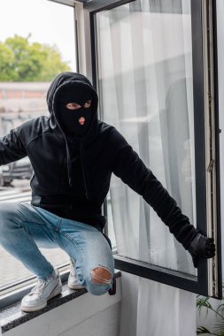 Robber in balaclava standing on open window during stealing clipart