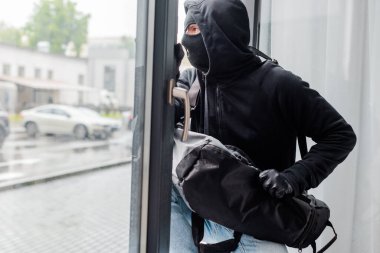 Robber in balaclava and leather gloves holding bag near open window  clipart