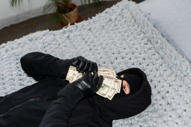 Robber in balaclava holding dollars near face while lying on bed  clipart