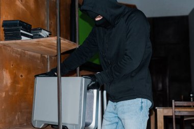 Robber in balaclava taking wireless speaker from cupboard during theft  clipart