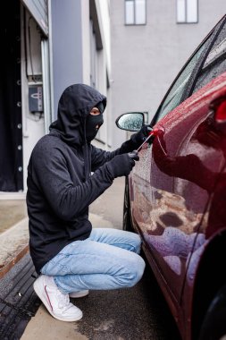 Sokak arabasının kapısını açarken tornavida kullanan kar maskeli soyguncunun yan görüntüsü 