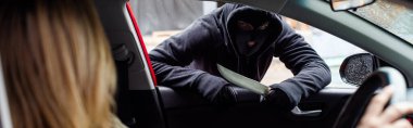 Panoramic shot of robber in balaclava holding knife near woman in car  clipart