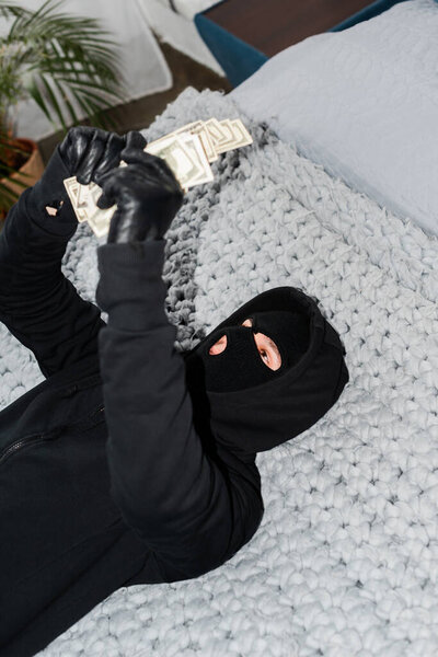 Selective focus of robber in balaclava holding dollar banknotes while lying on bed 