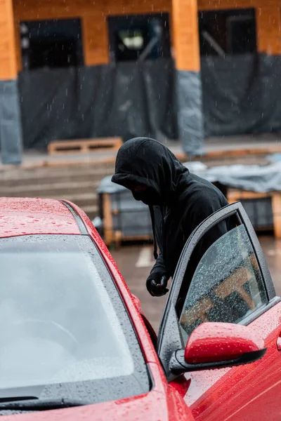 Side view of man in balaclava and leather glove standing near car with open door during robbery