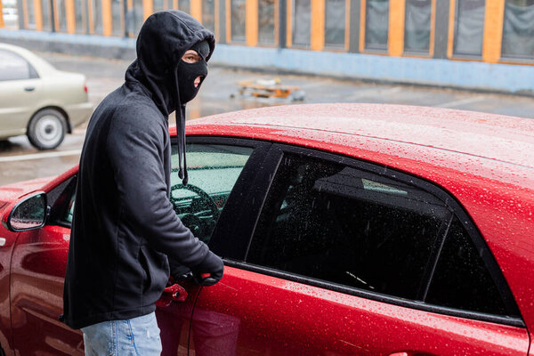 Грабитель в балаклавских и кожаных перчатках смотрит в сторону, открывая машину с отвёрткой на городской улице
 