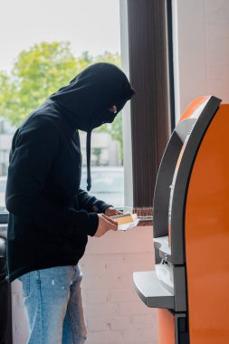 Kar maskeli soyguncunun ATM 'nin yanında kredi kartı tutarken yan görüntüsü 