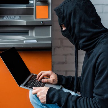 Side view of hacker in balaclava using laptop near automated teller machine clipart