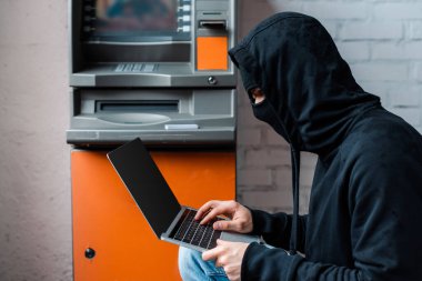 ATM 'ye yakın boş ekranlı dizüstü bilgisayar kullanan maskeli hacker yan görünümü 