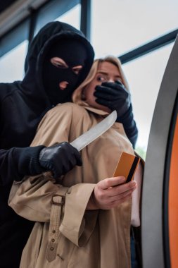 Düşük açılı bir bakış açısıyla soyguncunun elinde bıçak tutarken ağzı bankamatik yakınlarında kredi kartı tutan korkmuş bir kadına dönük. 
