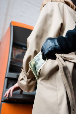 Cropped view of robber in leather glove taking dollar banknote from pocket of female coat near atm  clipart