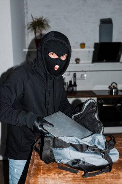 Kar maskeli bir soyguncu çantaya dizüstü bilgisayar koyarken kameraya bakıyor. 