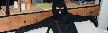 Panoramic shot of burglar in balaclava and leather gloves looking at camera on couch  clipart