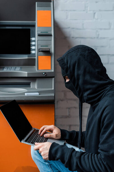 Side view of hacker in balaclava using laptop near atm 
