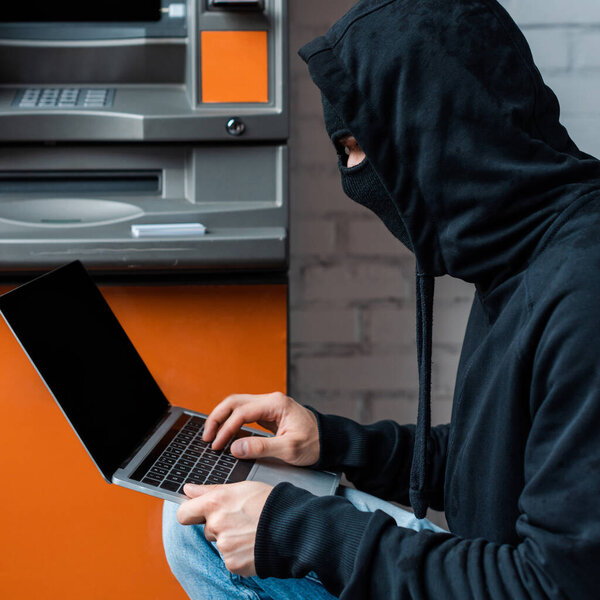 Side view of hacker in balaclava using laptop near automated teller machine