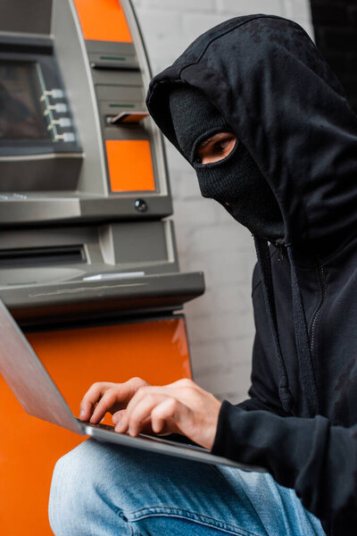 Side view of thief in balaclava using laptop near automated teller machine