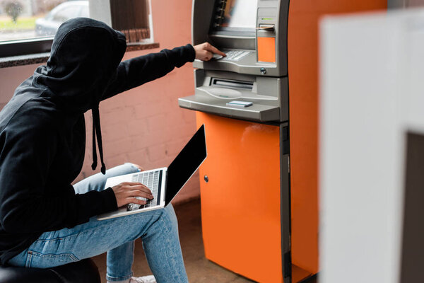 Side view of hacker in mask using atm and laptop with blank screen 