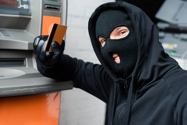 Smiling thief in balaclava looking at camera while holding credit card near atm  